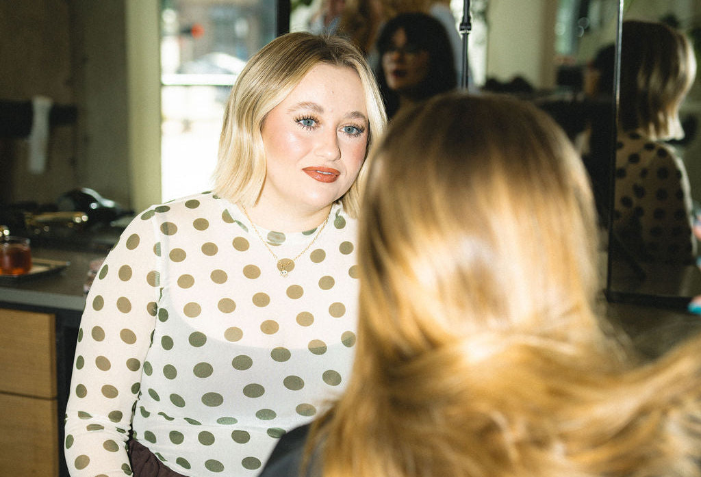A woman with blonde hair wearing a cream long-sleeve top with brown polka dots sits in a salon chair facing another woman with long blonde hair in the foreground, with salon mirrors and a window visible in the background.