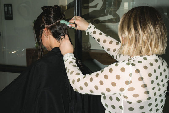 A woman with blonde hair sits in a salon chair while another woman styles her hair. The stylist wears a sheer white long-sleeve top with brown polka dots and dark brown pants, while the client wears a black salon cape.
